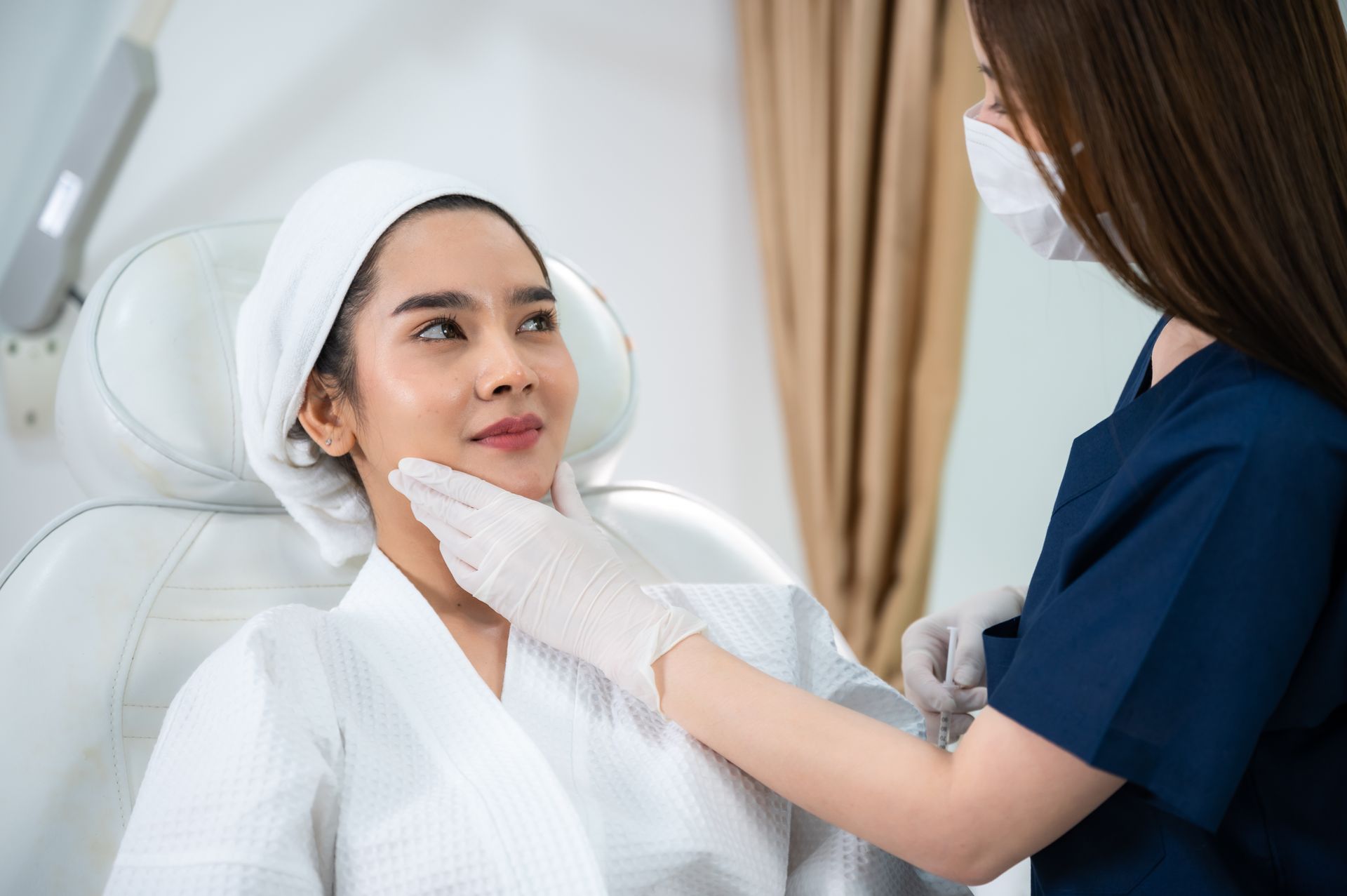 A professional in a mask and uniform examines a patient’s face in a clinical spa setting.