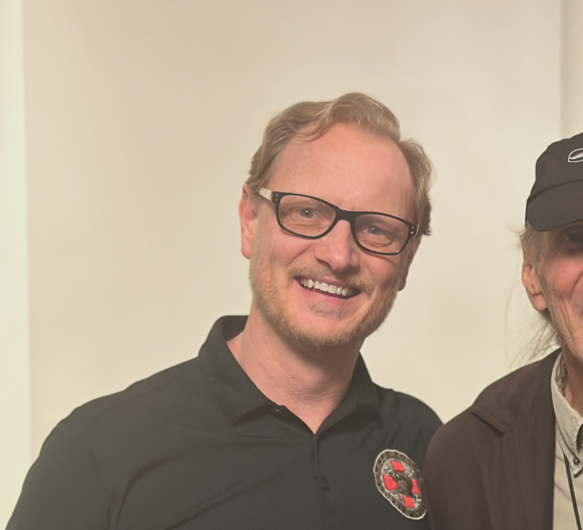 A man with a beard is wearing a red polo shirt and smiling.