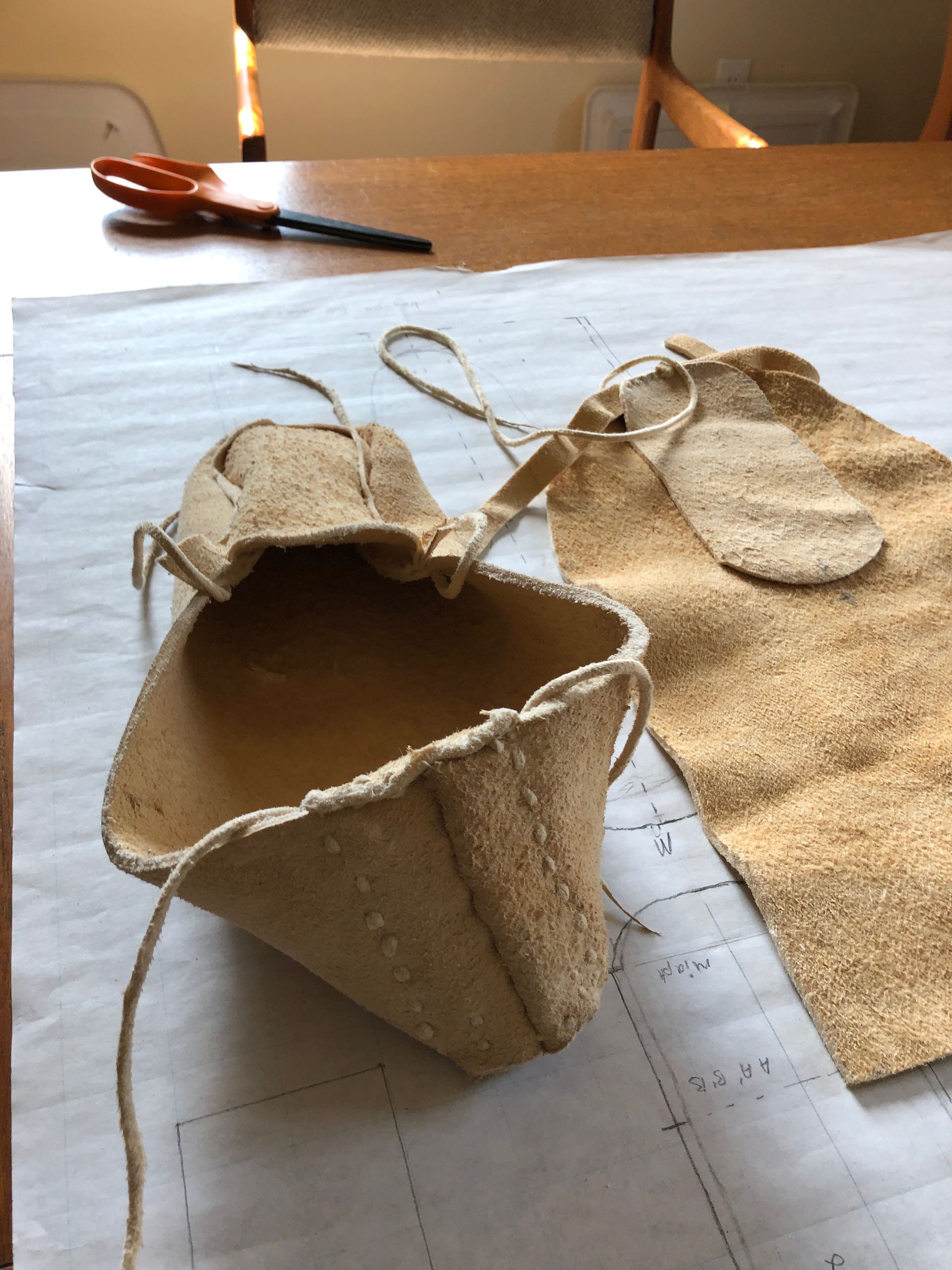Tan leather pouch with drawstring, partially constructed on a paper surface, scissors in background.