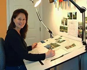 Woman at a drafting table smiles while working. The table is lit by two lamps and has photos pinned above.