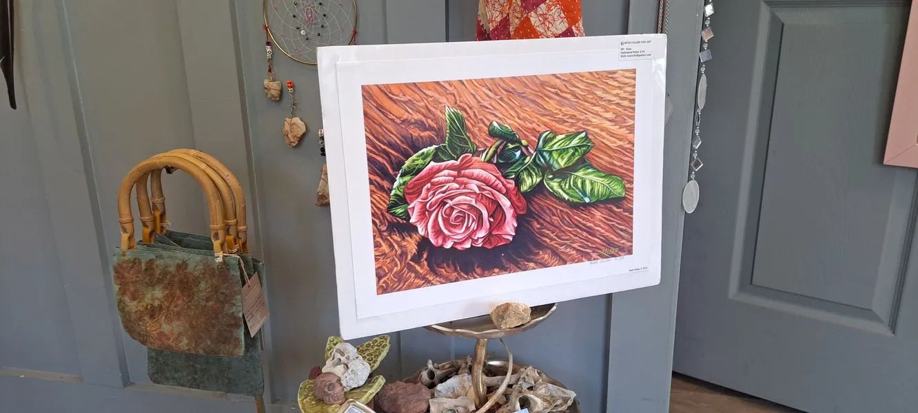Painting of a pink rose on a wooden surface, displayed on a stand.  An artisanal bag hangs to the left.