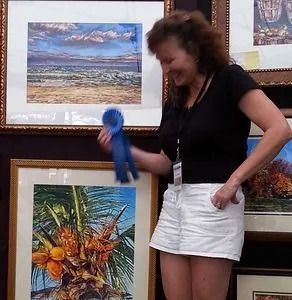 Woman holding blue ribbon, smiling, next to framed art. She wears a black shirt and white shorts.
