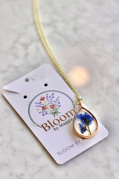 Gold necklace with blue flower pendant on a tag for Bloom by Maggie.