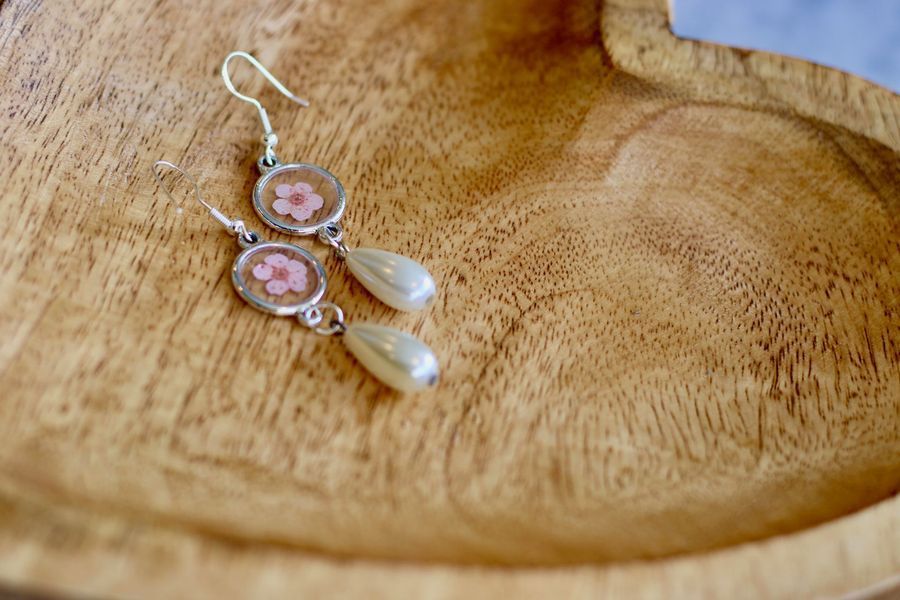 Dangling earrings with pink flower and pearl accents on a wooden heart-shaped tray.