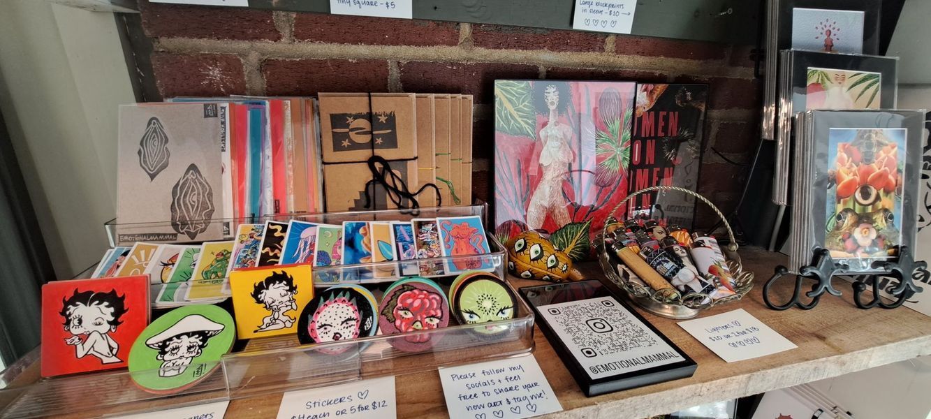 A display of artwork, including paintings, coasters, and small framed pieces, arranged on a wooden shelf in a shop.