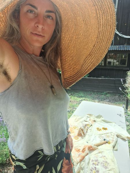 Woman wearing oversized hat, posing outdoors, armpit hair visible. A garment lies on a nearby table.
