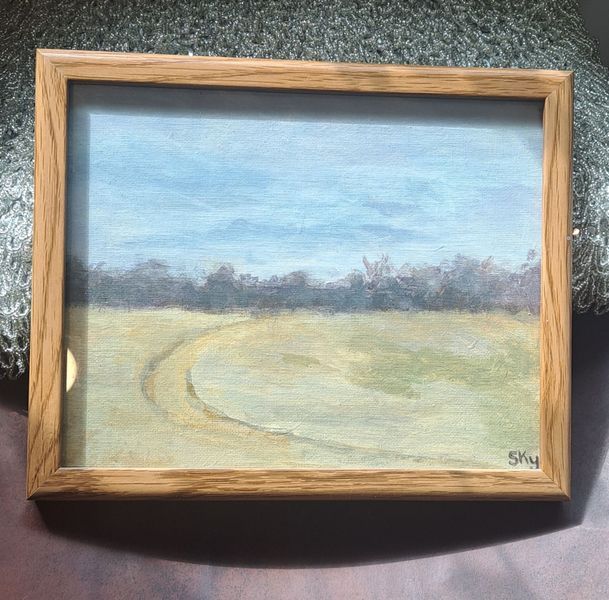 Landscape painting of a field with a curved path, trees, and cloudy sky. Framed in light wood.