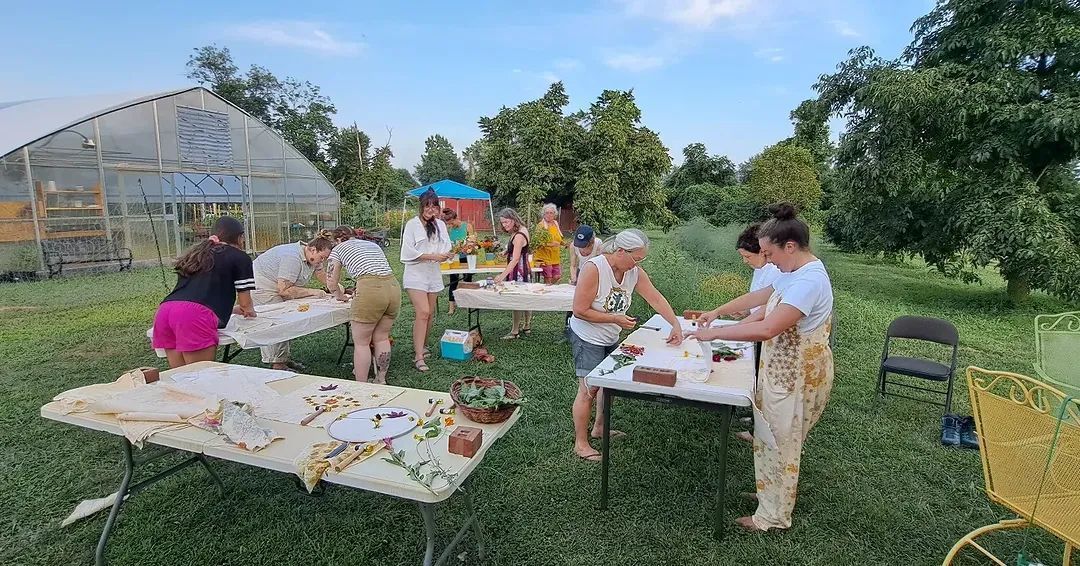 People crafting at tables outdoors, greenhouse and trees in background.
