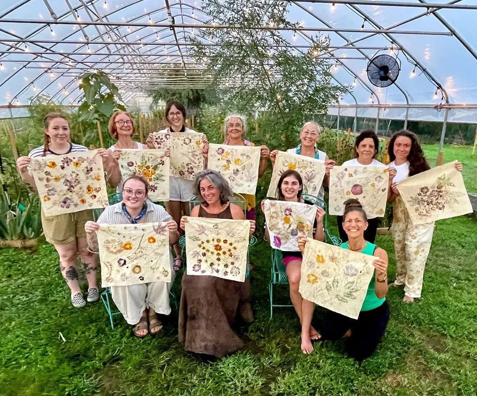 Group of people holding up decorated cloths, smiling in a greenhouse.