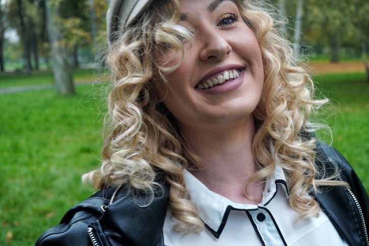 Woman with curly blonde hair smiles outdoors, wearing a white shirt, black jacket, and cap.