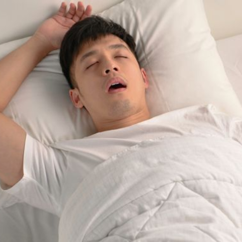 Man sleeping in bed, mouth open, hand raised. White sheets and pillow.