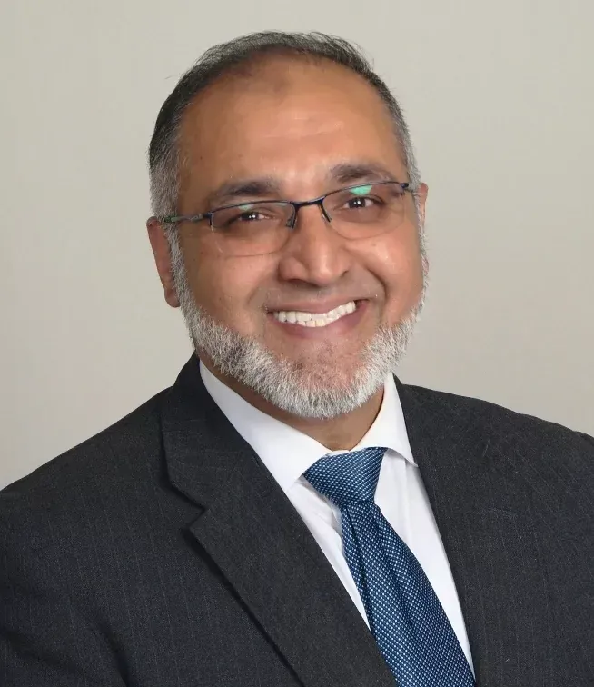 Man in suit and glasses smiles at the camera; neutral background.