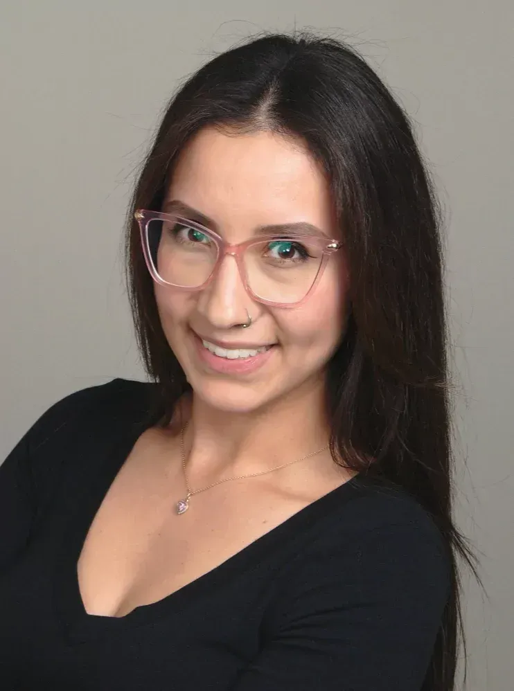 Woman with long dark hair wearing pink-rimmed glasses smiles, small nose ring, black top.