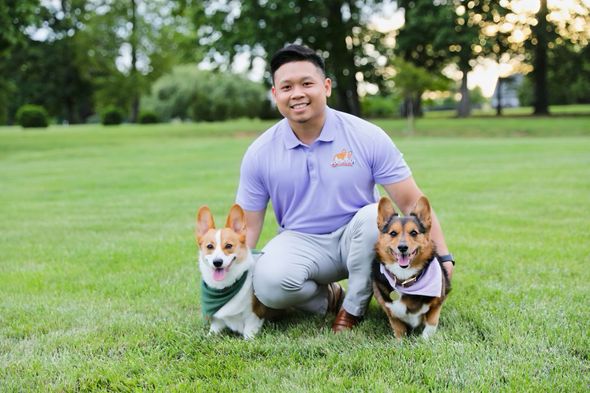 A man is kneeling down with two corgi dogs in a park.