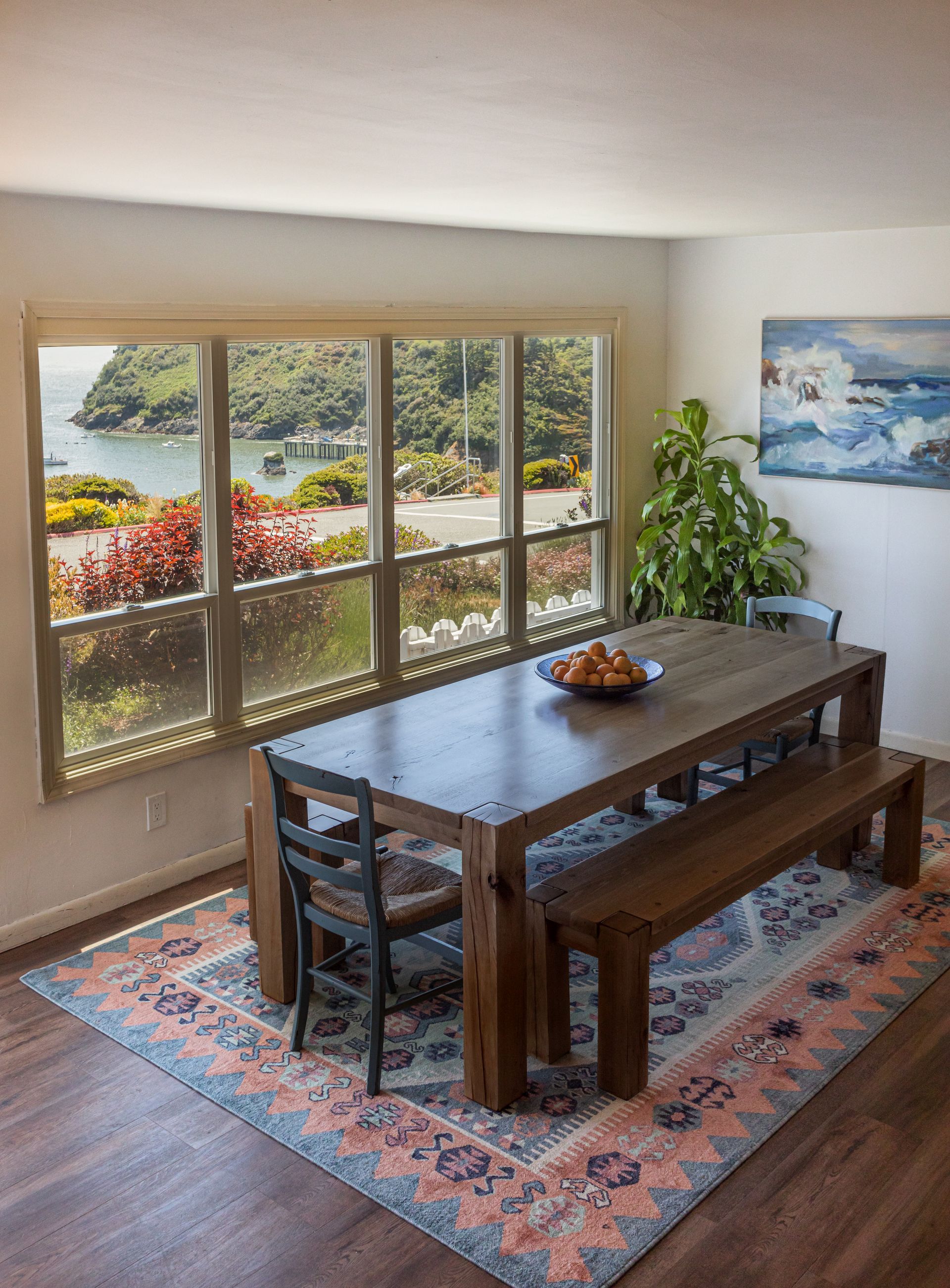Dining room with wooden table, benches, and chair. Large window with harbor view, colorful rug on hardwood floor.
