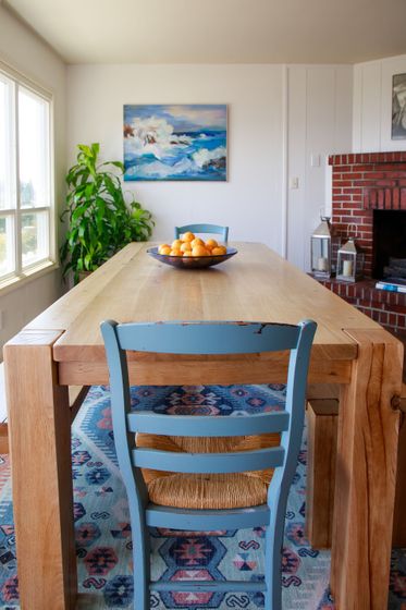 A cat is sitting on a chair in front of a wooden table with a bowl of oranges on it.