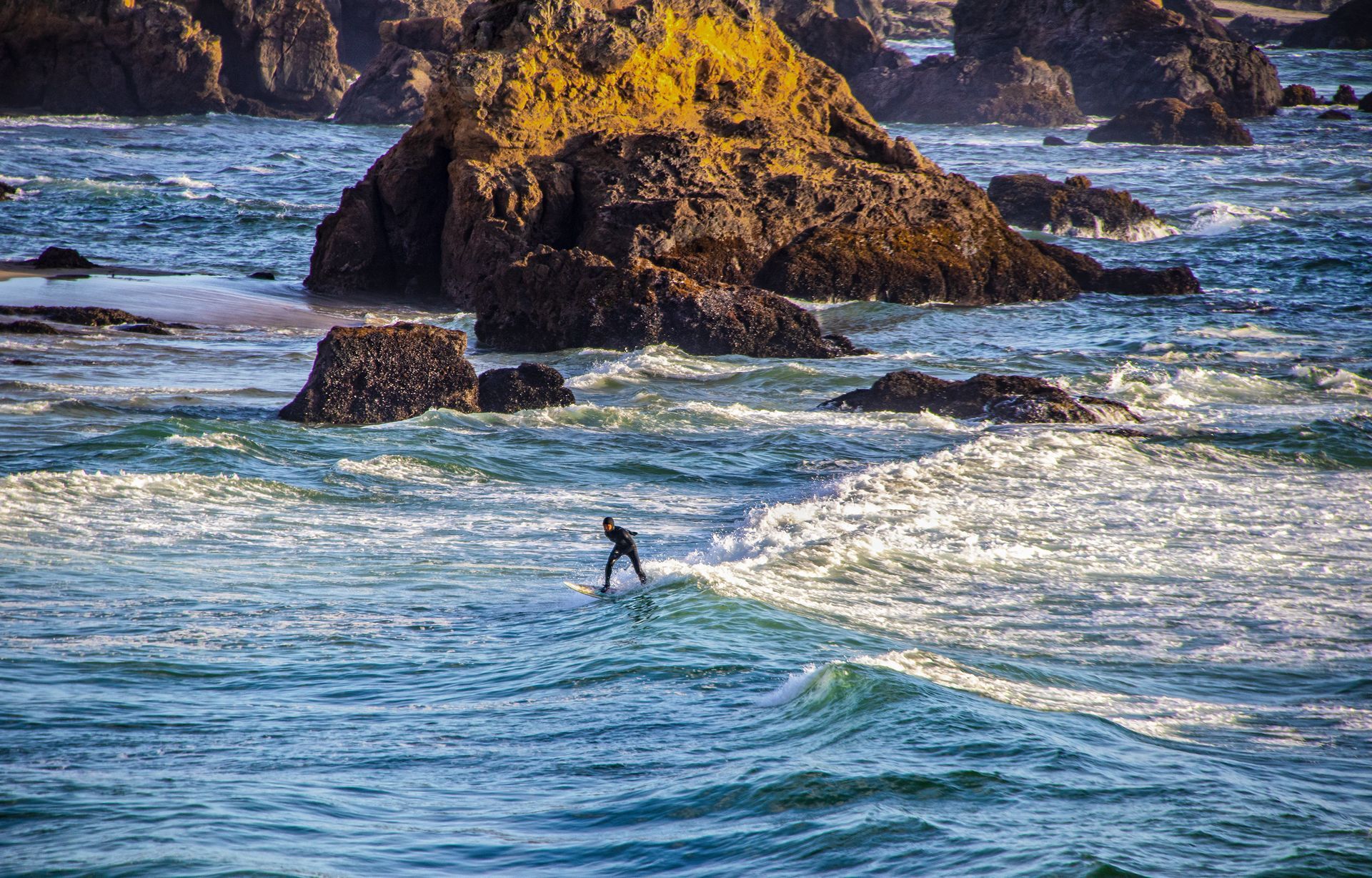 A surfer in a wetsuit rides a small wave near large, rocky formations in a coastal ocean setting.
