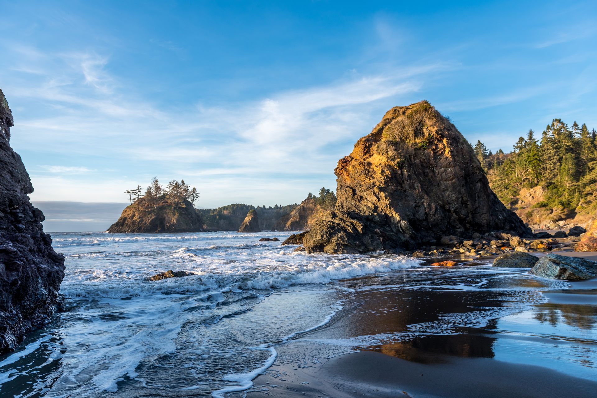 A rocky coastline with large sea stacks and trees under a blue sky, with gentle waves washing onto the sandy beach.