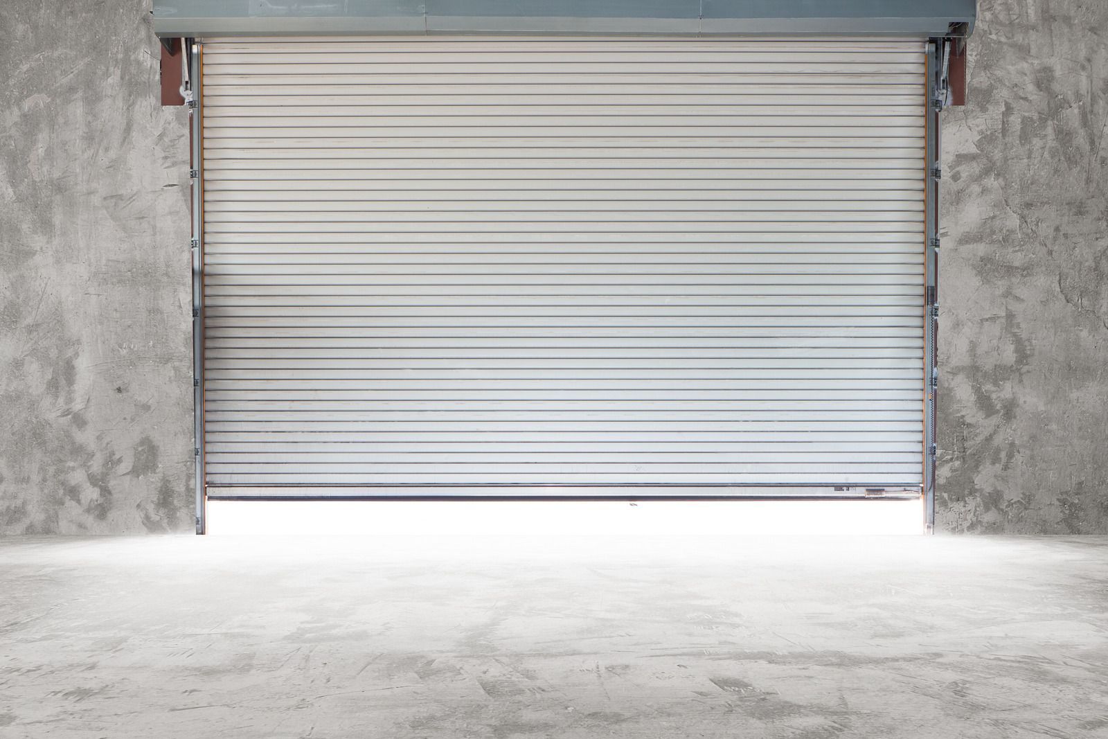 Closed metal roll-up door in concrete wall opening, casting a bright light onto a concrete floor.