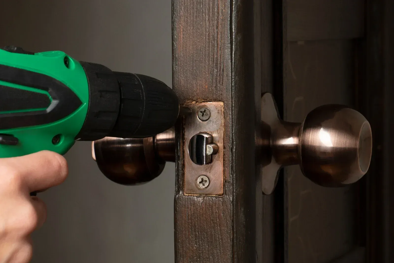A person drilling a door lock with a green power drill, presumably to remove or replace it.