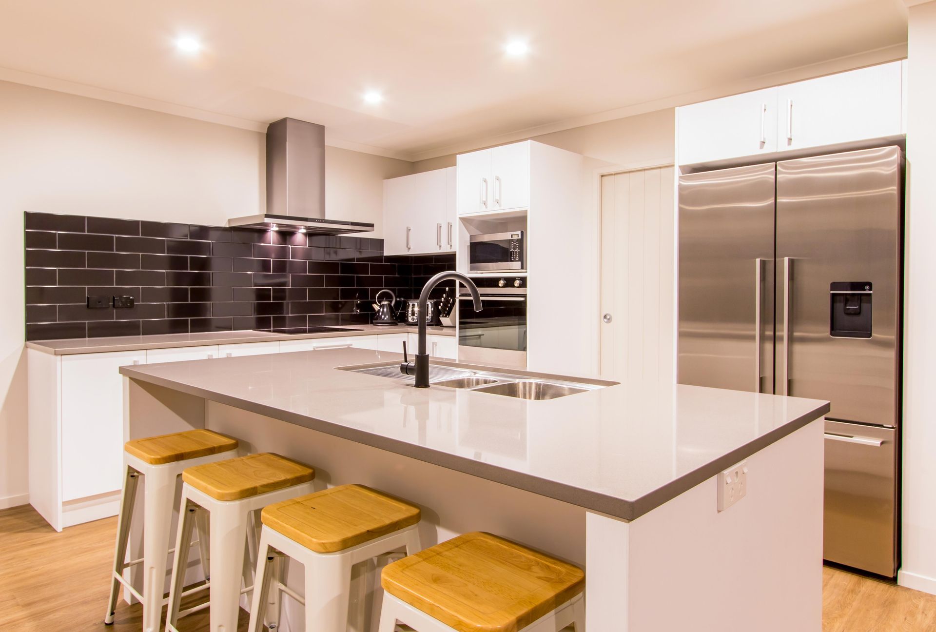 A kitchen with stone benchtops, a tiled black splashback, a black tap, and wood flooring.