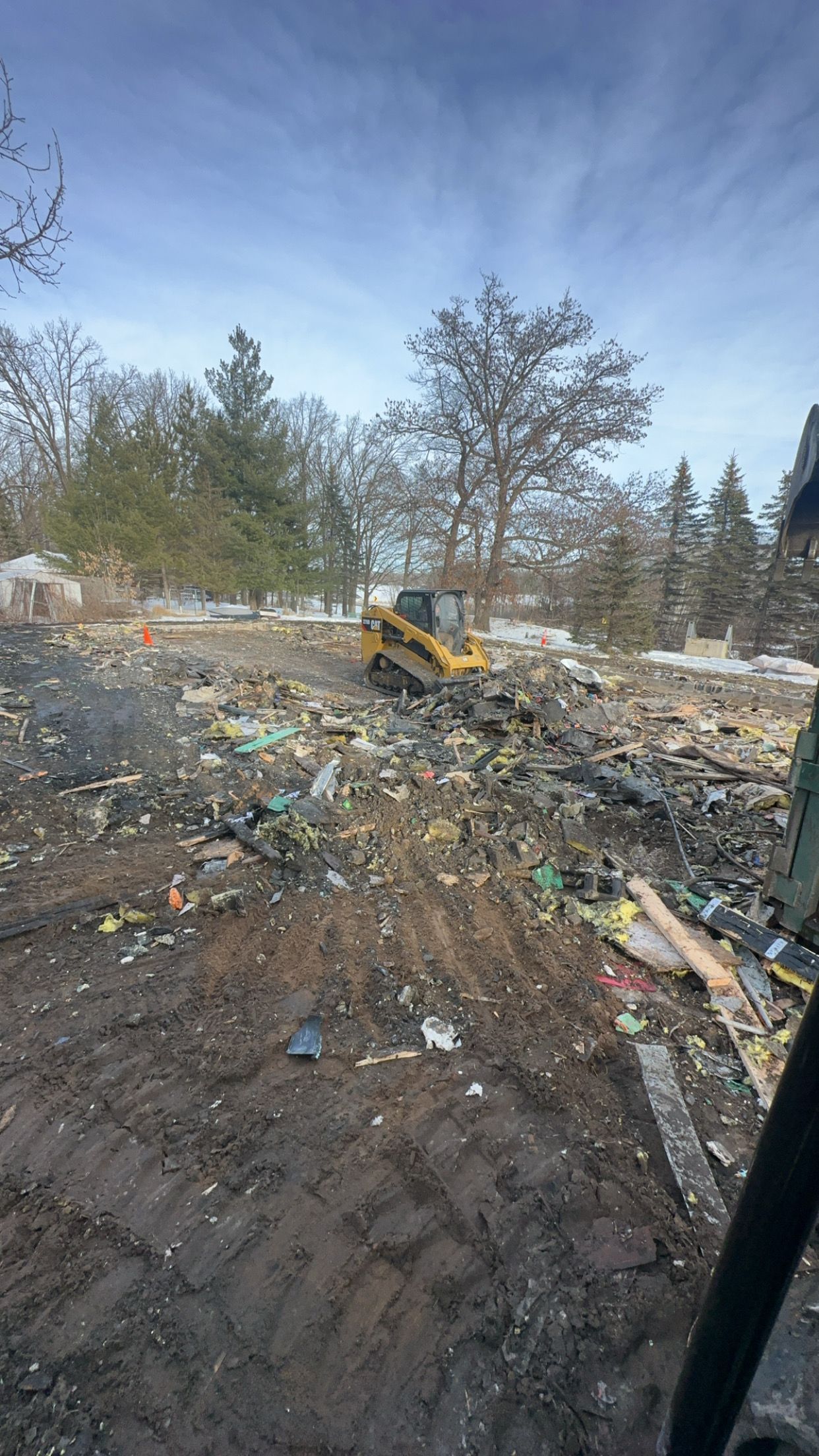 A yellow skid steer clears a debris-filled, muddy construction site under a partly cloudy sky.