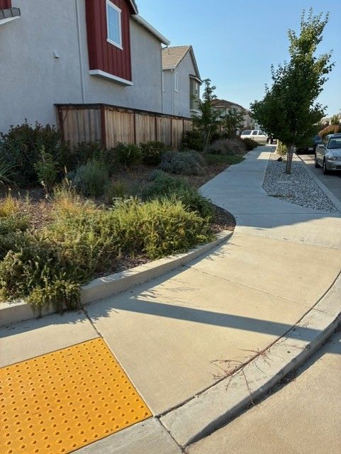 Cleanup King LLC - Sidewalk curving past landscaping and a wooden fence, with a yellow tactile tile and a red-accented building.