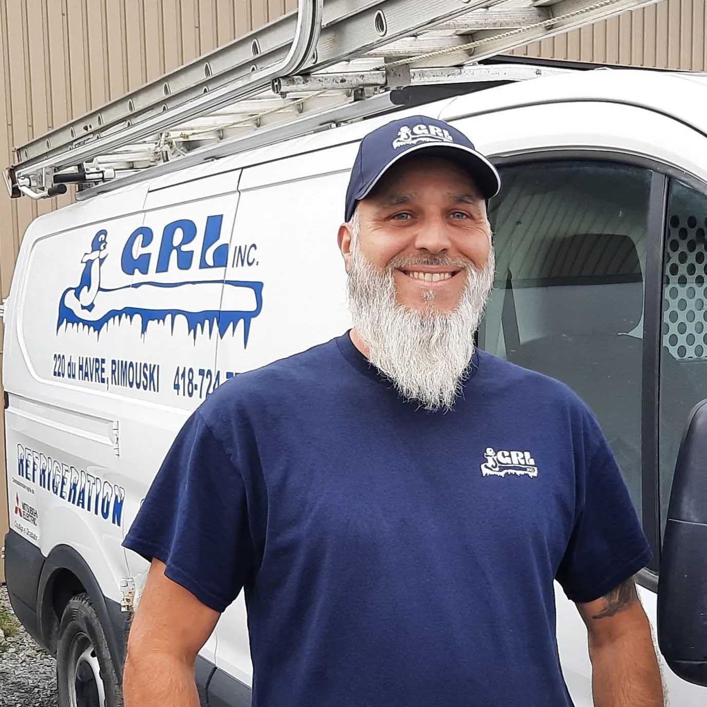 Un homme barbu, vêtu d'une chemise et d'une casquette bleu marine, sourit devant une camionnette de l'entreprise avec une échelle et le logo « GRL Inc. »