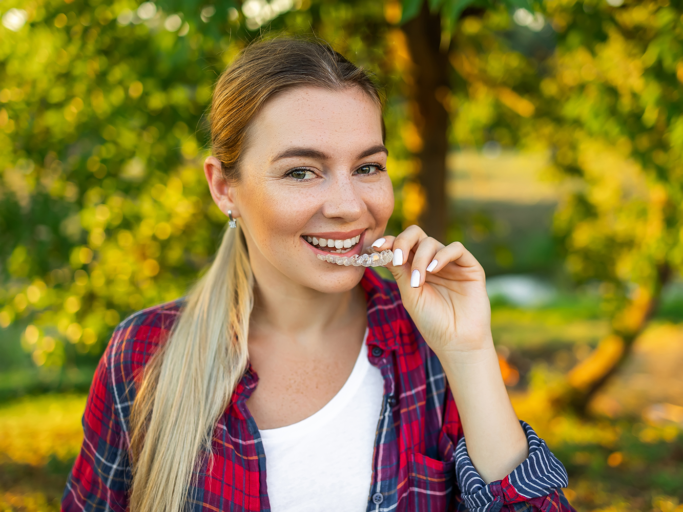 girl standing outside smiling holding a clear aligner tray and smiling while placing the tray in her mouth