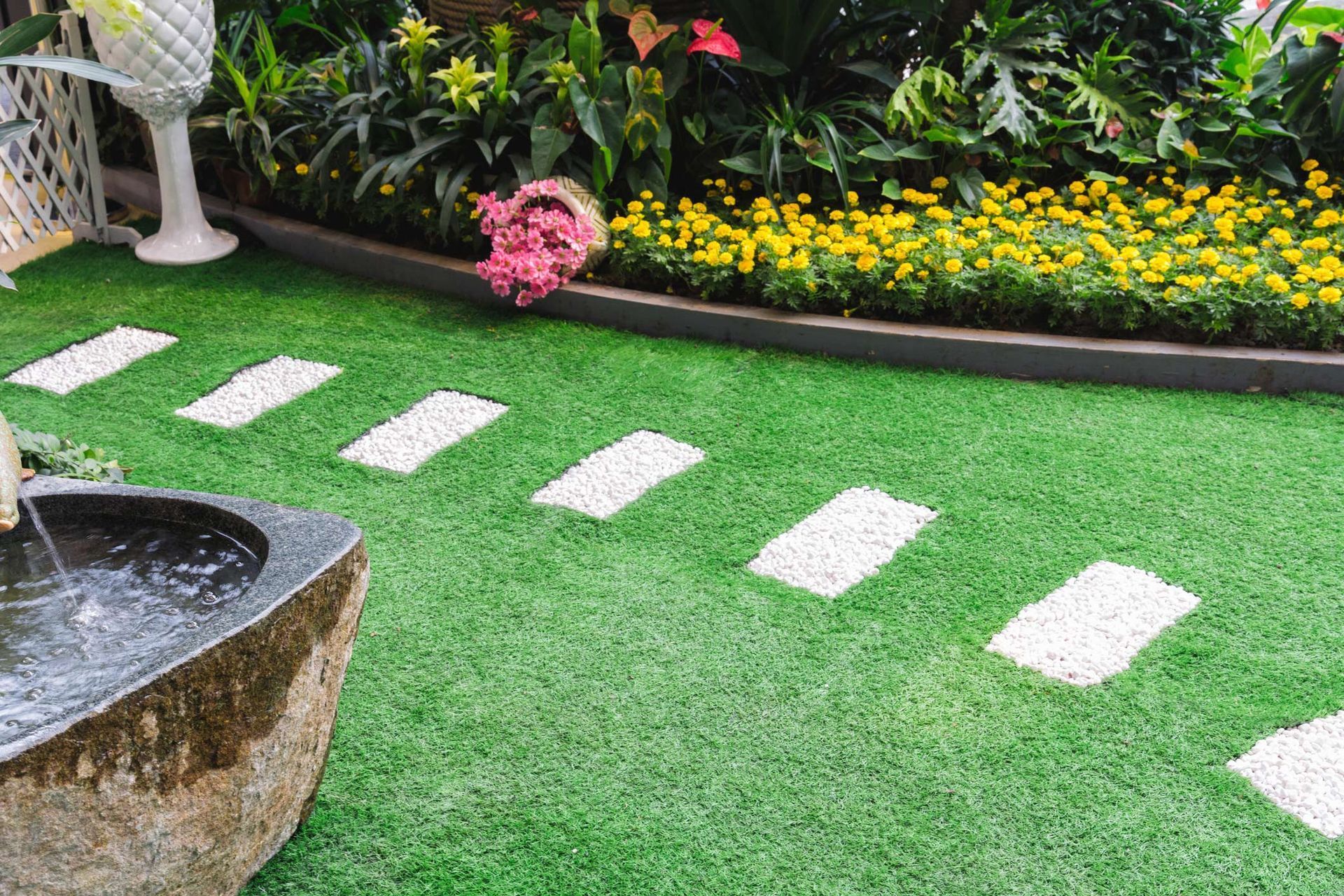 A stone water feature sits on artificial turf next to a path of white stone stepping stones leading to a flower bed.