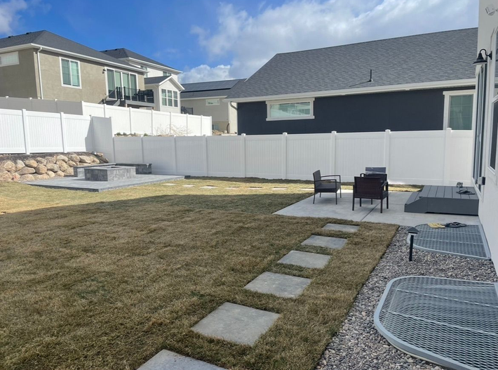 Backyard patio with stone path, lawn, white fence, and outdoor chairs under a blue sky Backyard patio with stone path, lawn, white fence, and outdoor chairs under a blue sky