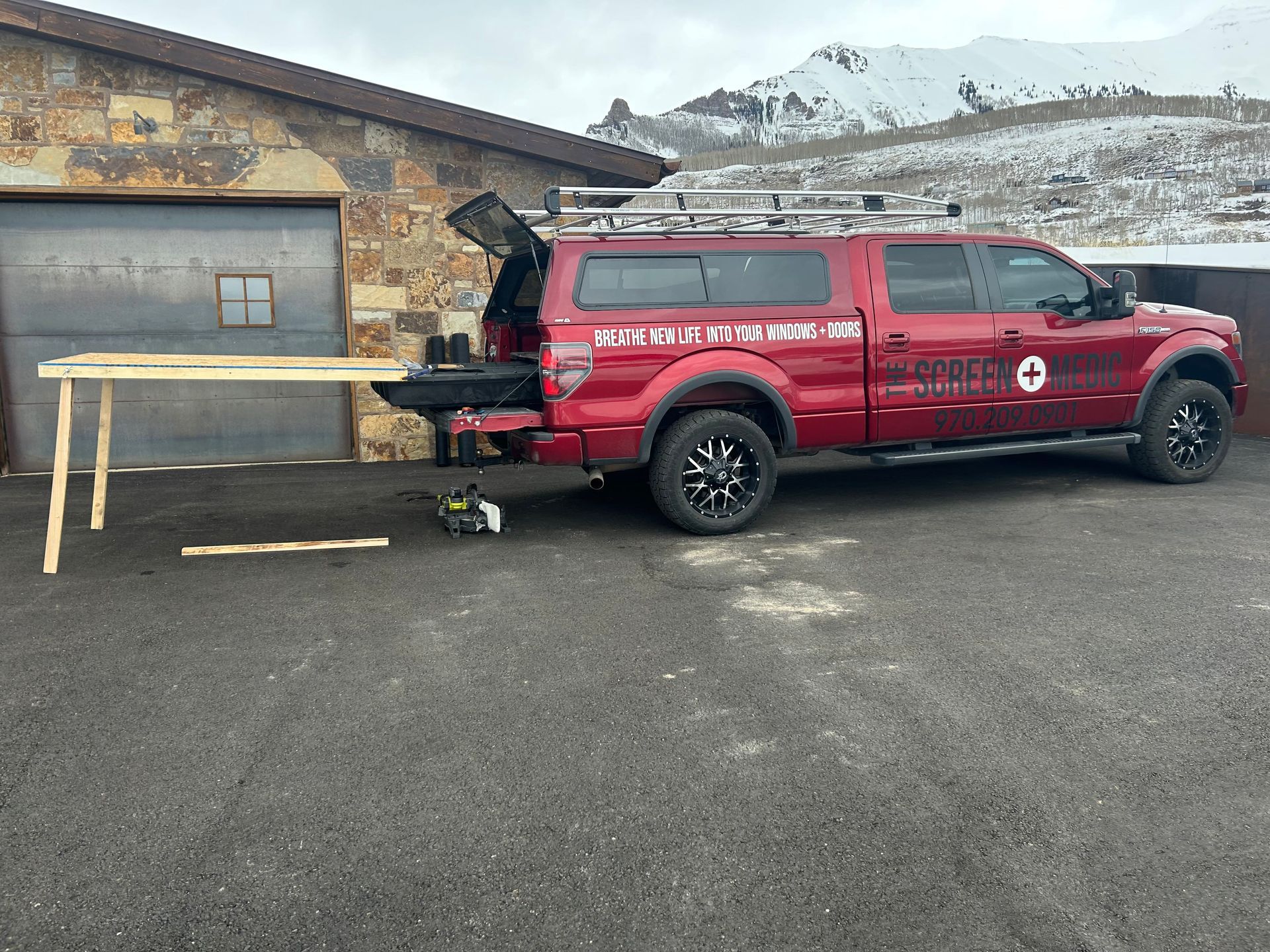 A red truck is parked in front of a garage with a wooden table in the back.