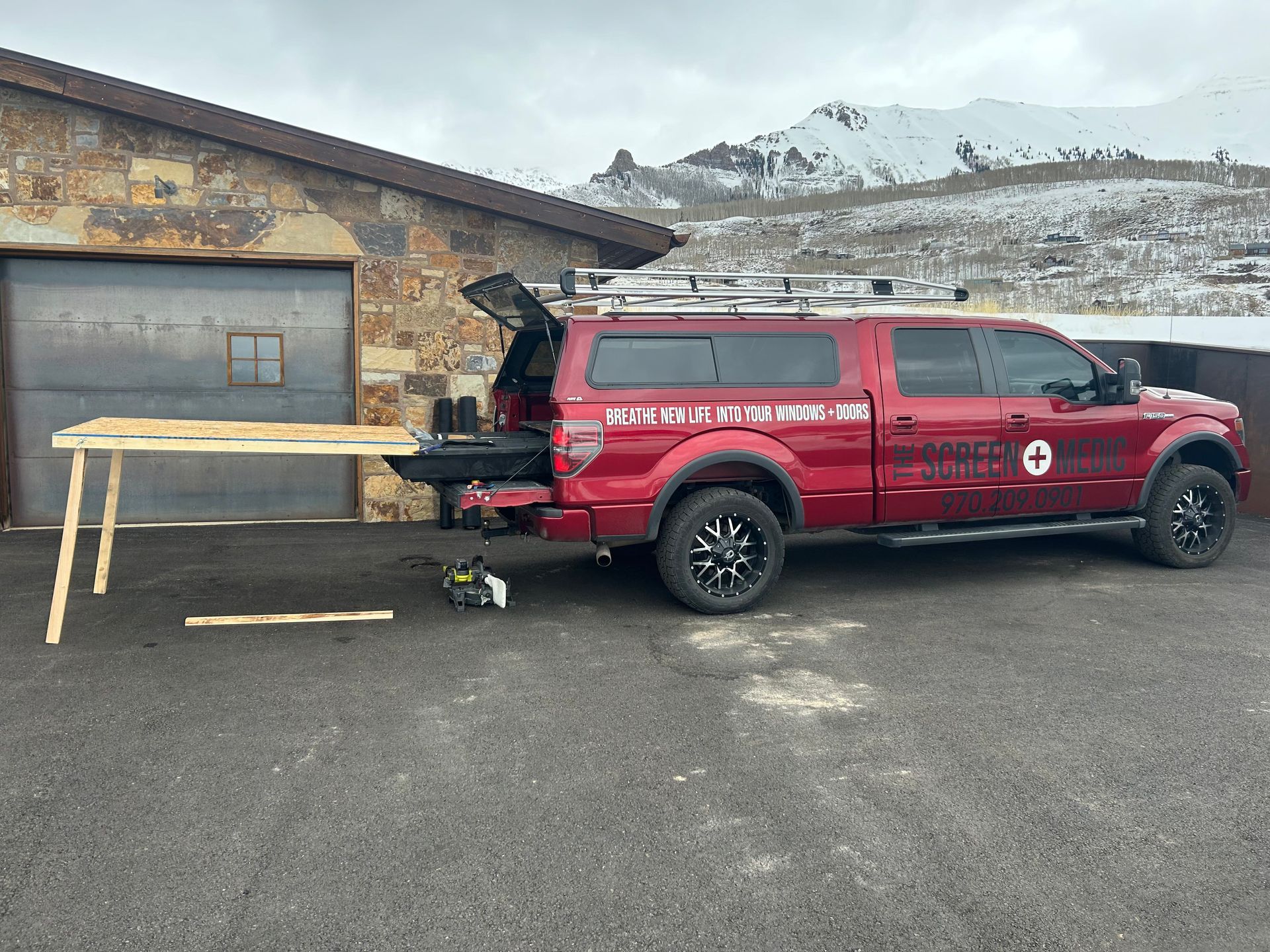 A red truck with a wooden table attached to the back is parked in front of a garage.