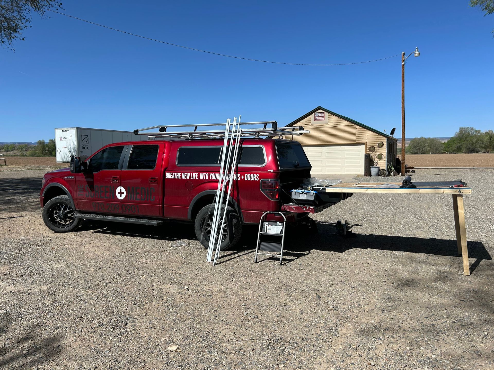 A red truck with a ladder attached to the back is parked in a gravel lot.