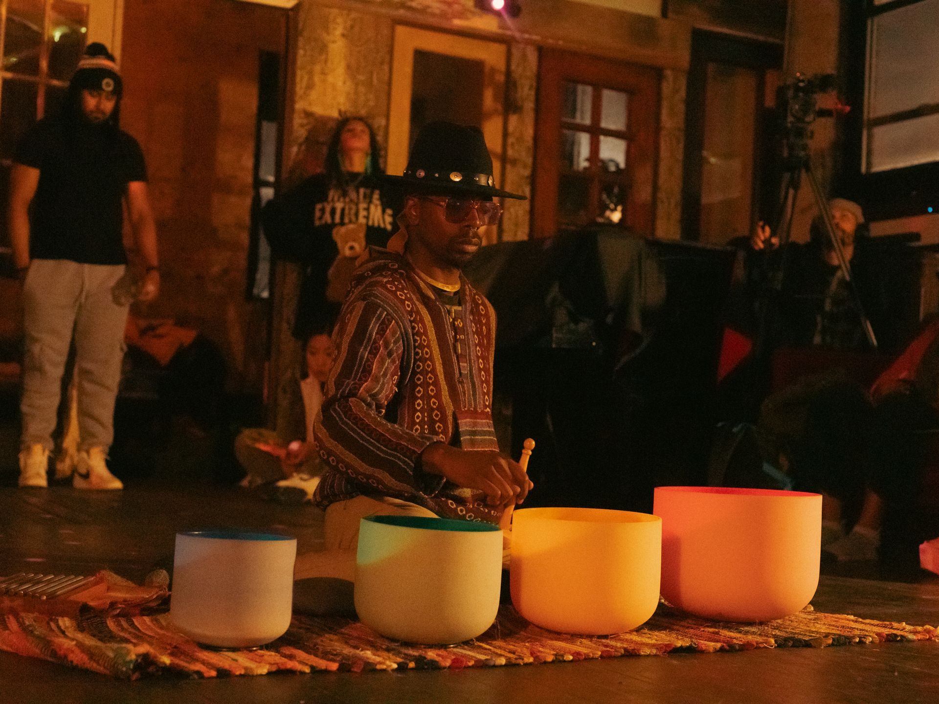 Man playing crystal singing bowls, outdoors at night. Others watch.
