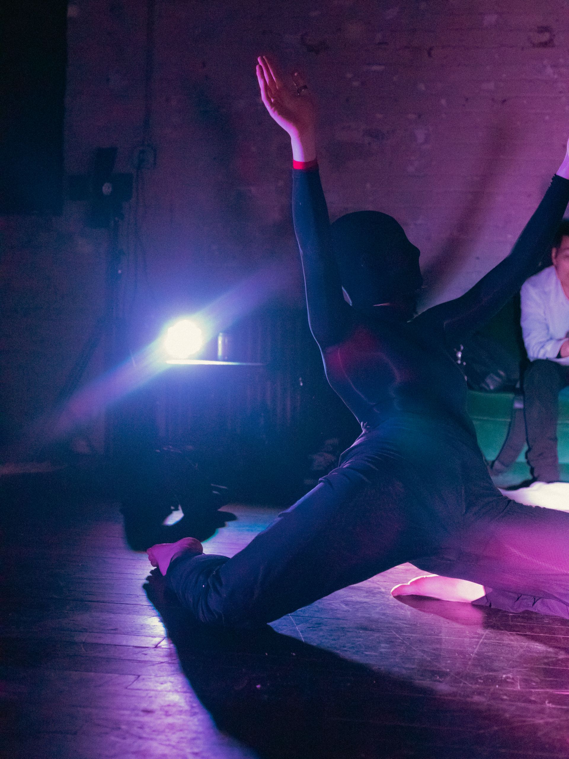 Dancer in black leans on one knee, arms raised. Stage lit with purple and white light.