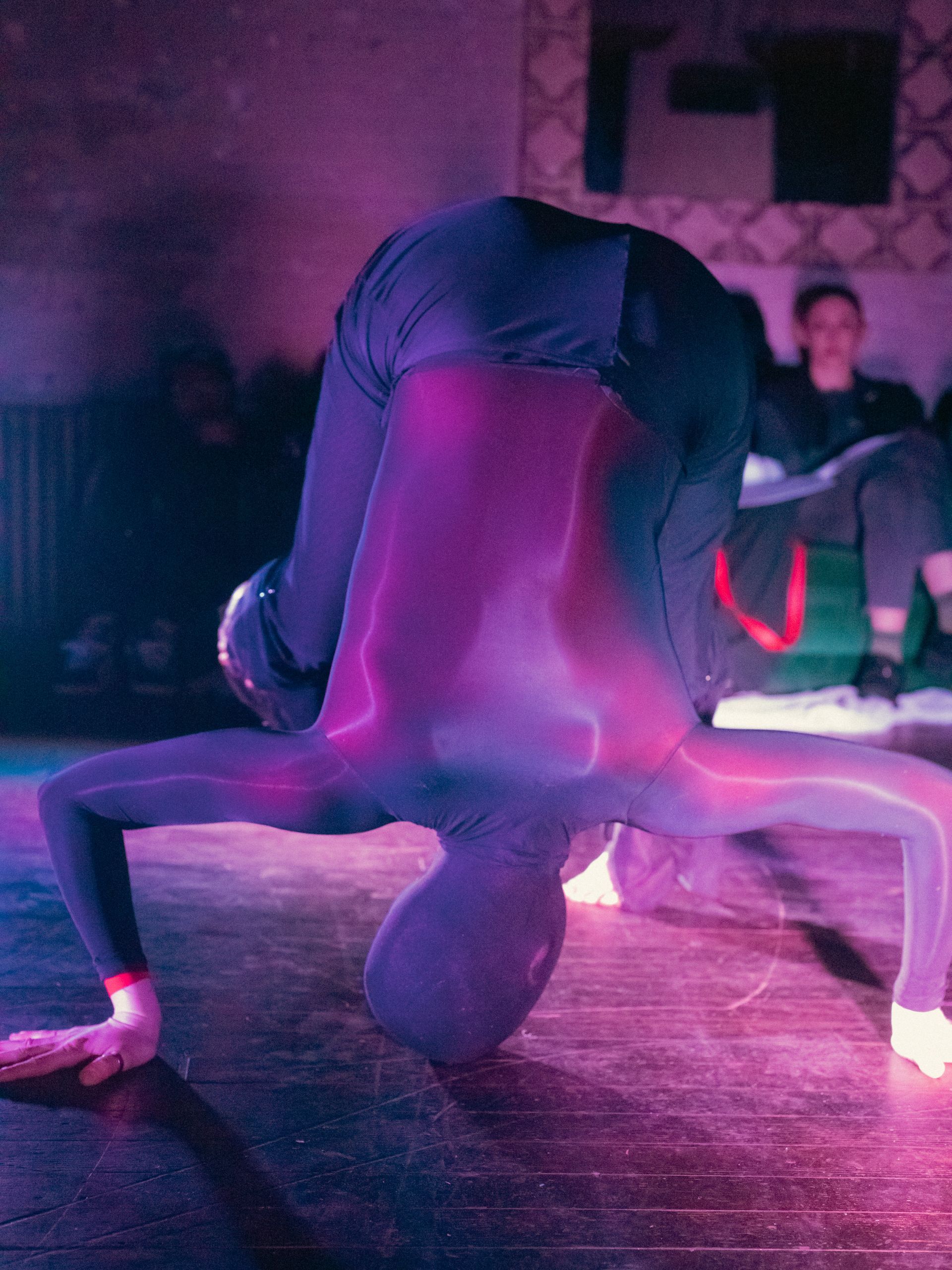 A person in a black bodysuit doing a headstand on a wooden floor, with a purple glow effect.