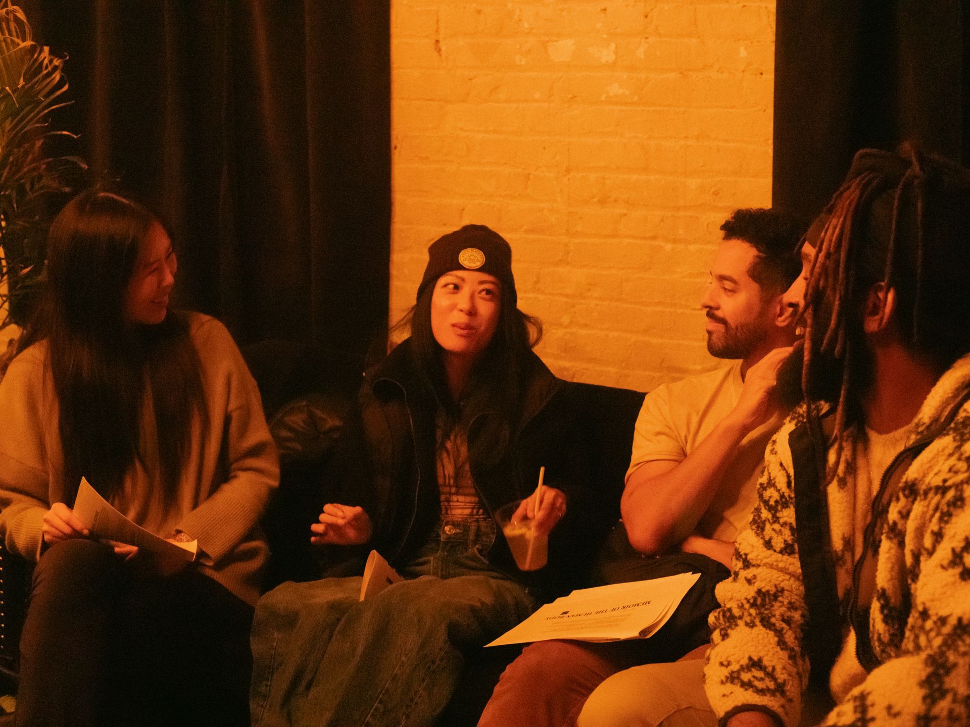 Four people in a dimly lit room, engaged in conversation. One woman gestures, others listen, papers visible.