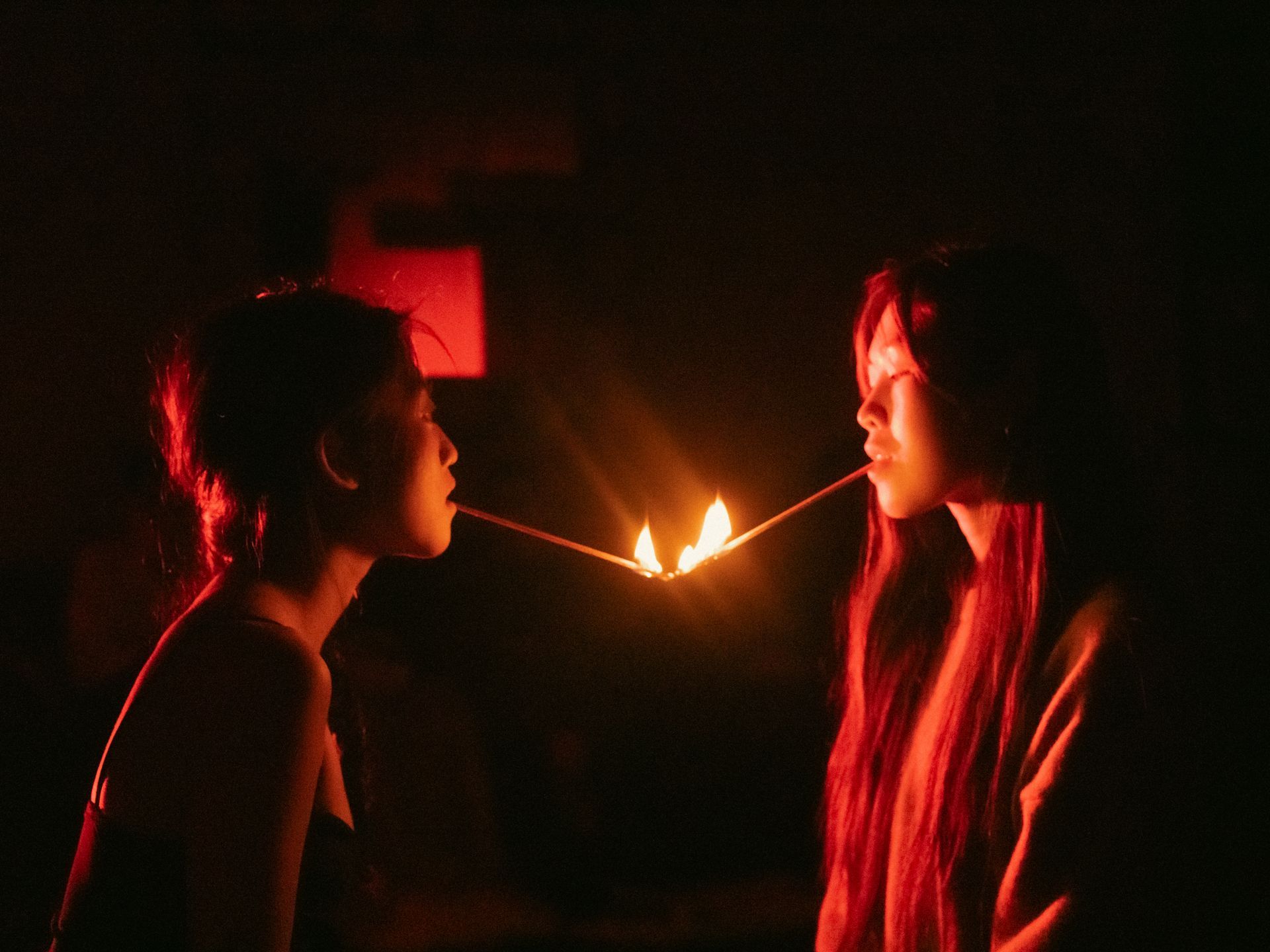 Two women with matchsticks between their mouths, flames glowing. Dark setting, red lighting.