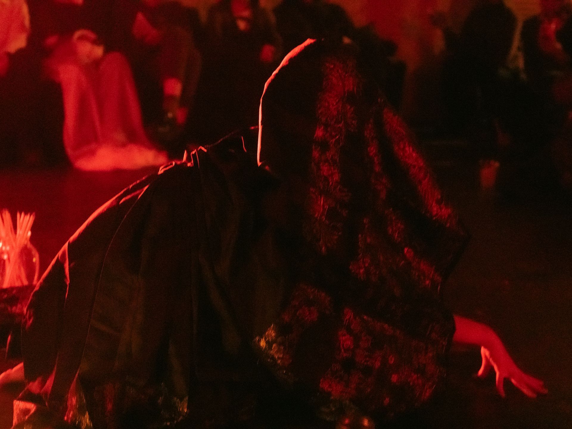 A person in dark clothing and lace veil, possibly dancing, in a dimly lit space with others. Red lighting.