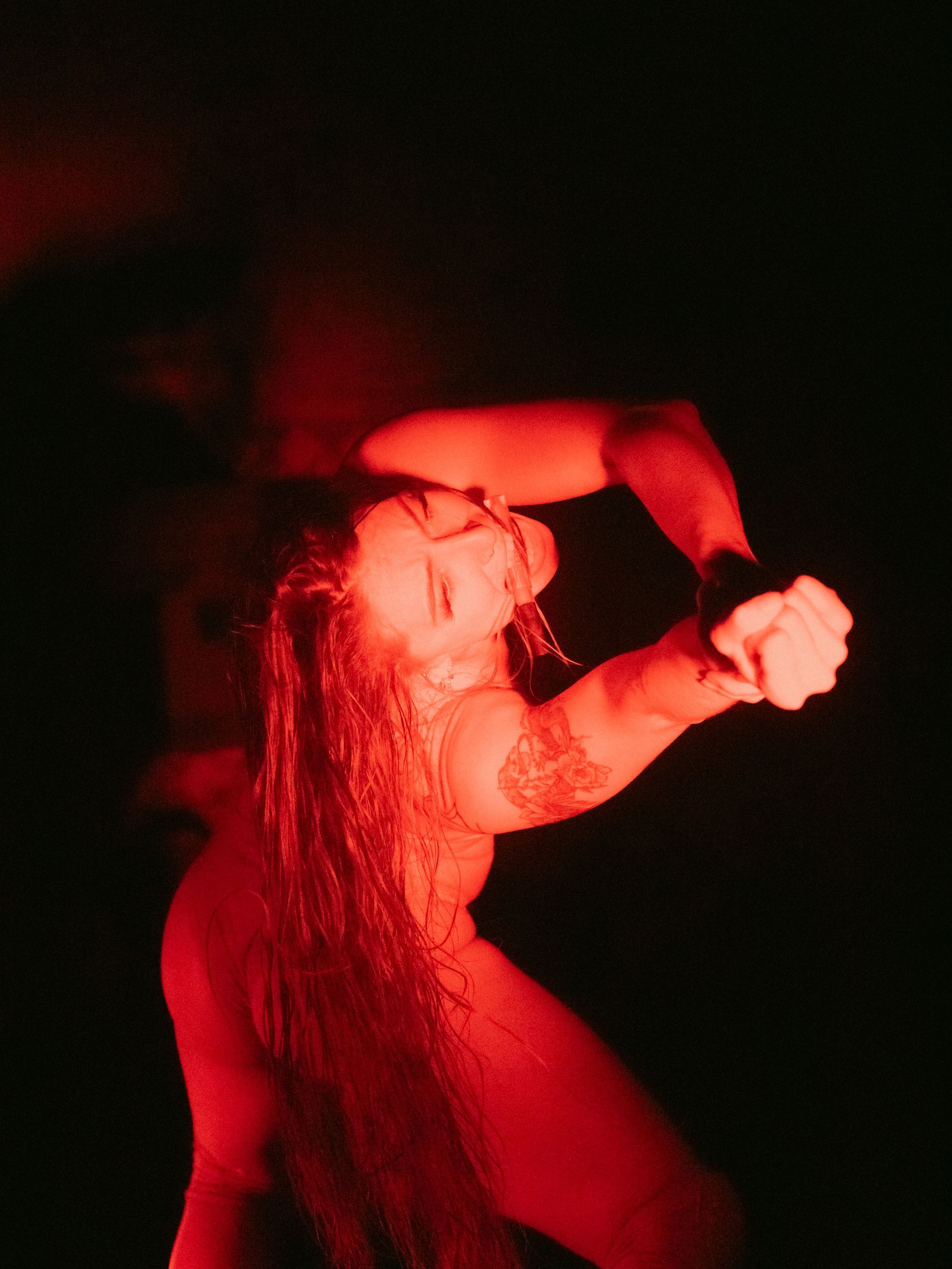 Woman in red light, arms raised, head tilted back, fist clenched.