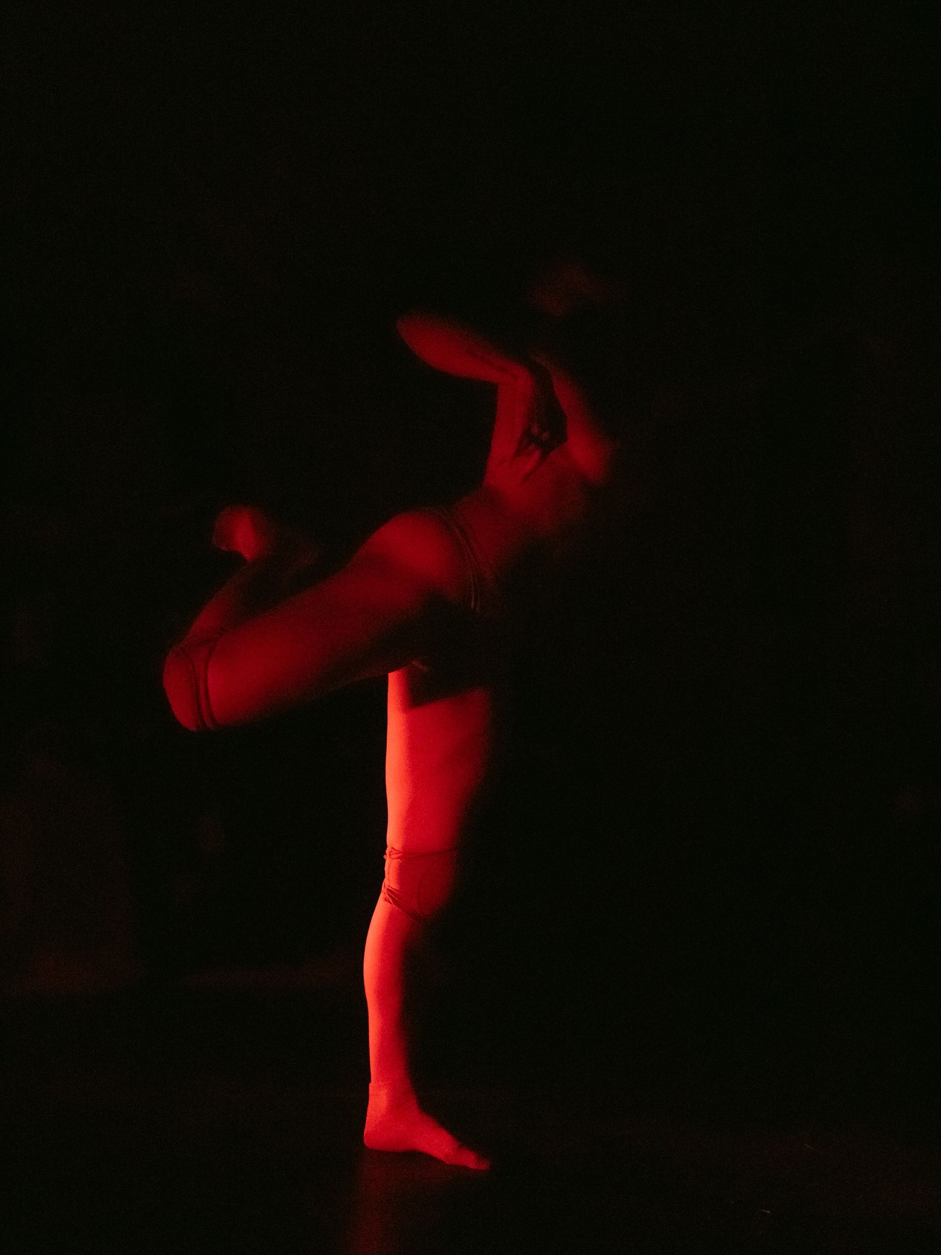 A dancer in red light performing a ballet move on a dark stage.