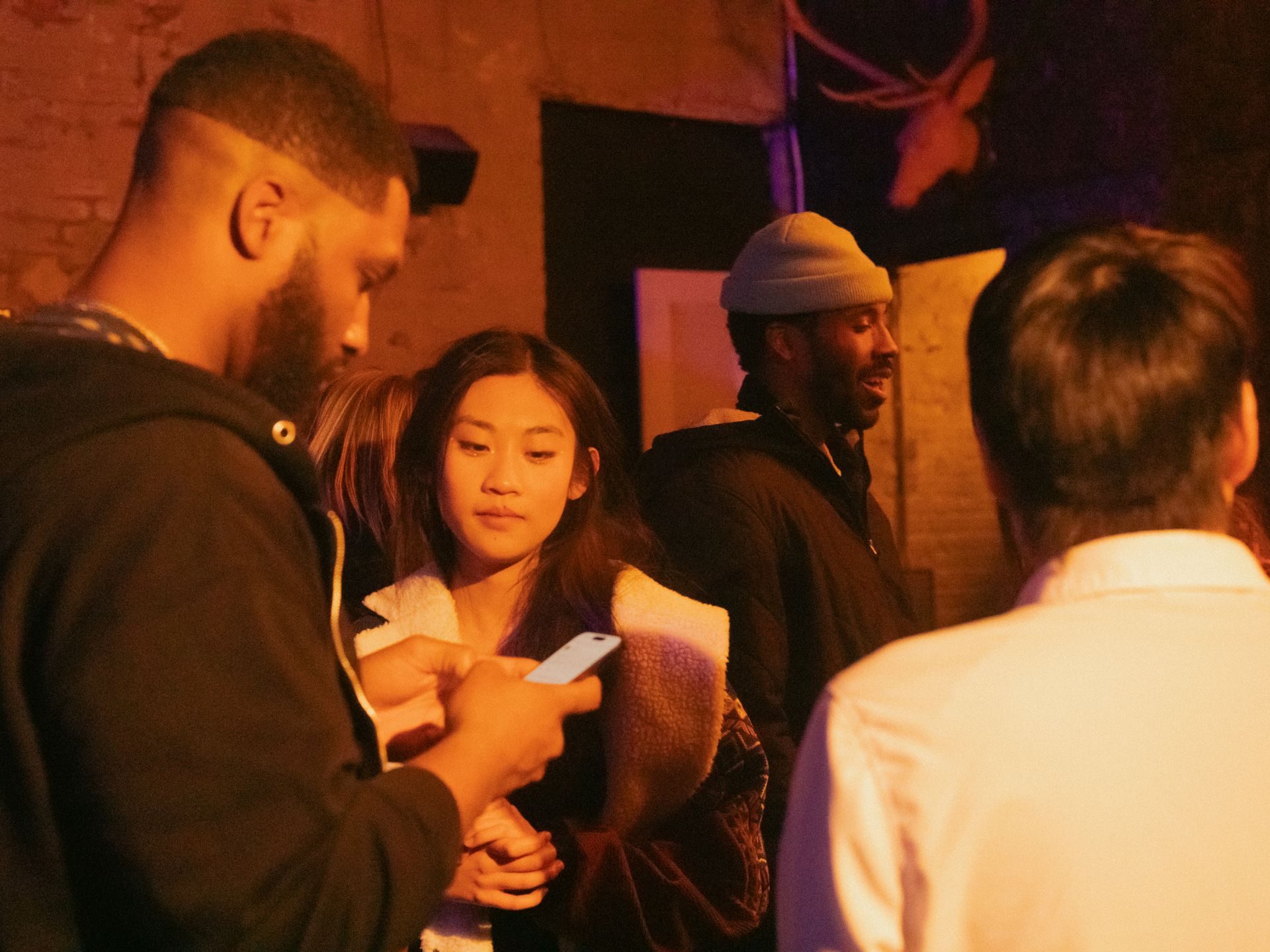 A group of people are gathered in a dimly lit space. A woman checks her phone, as three men look on.