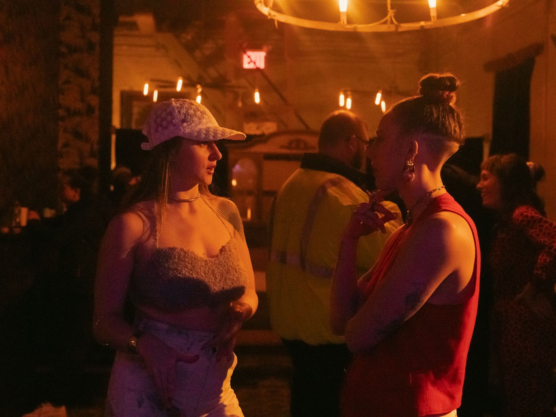 Two women conversing at a dimly lit event; one wears a hat, the other has a shaved head.