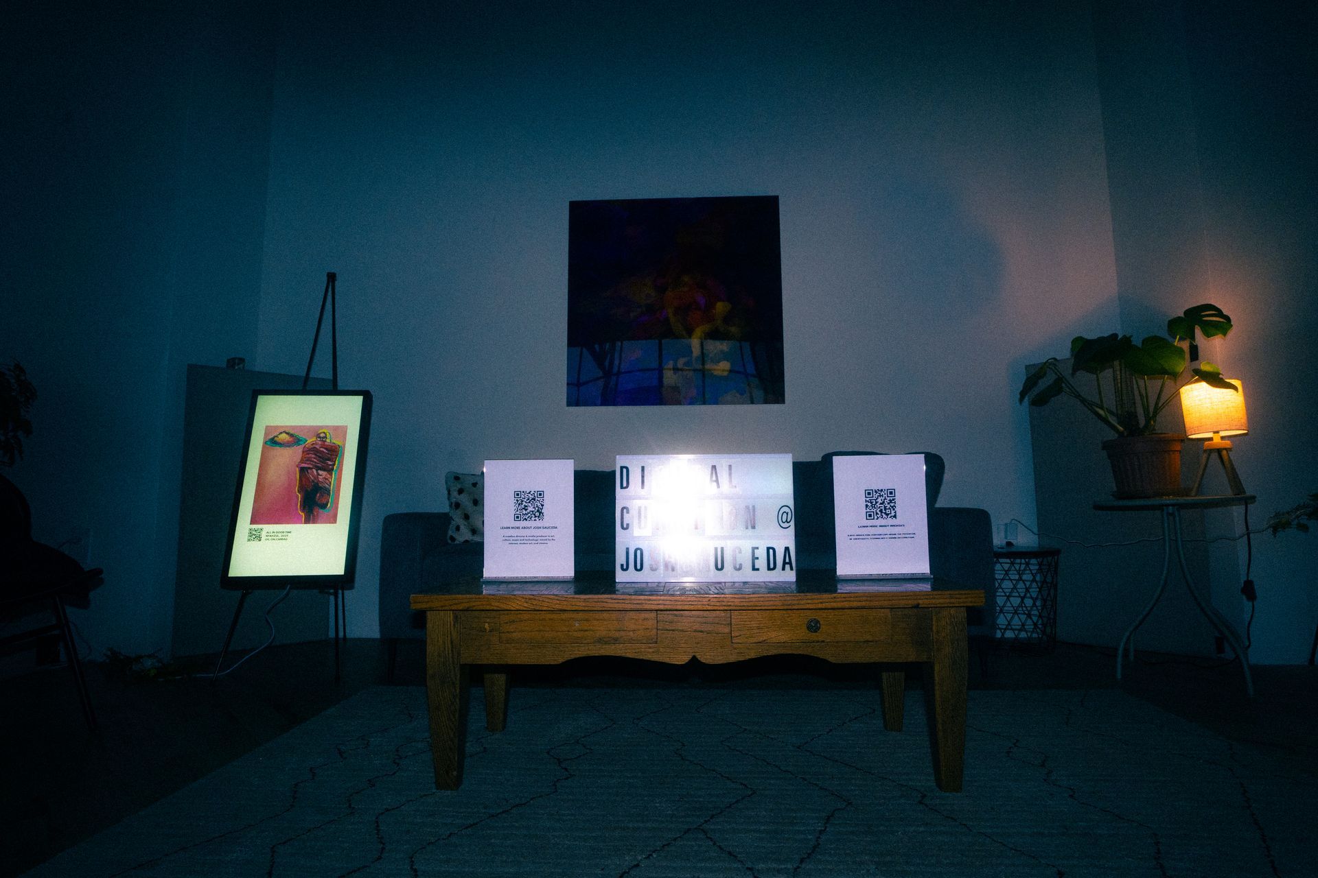 Dark room with coffee table displaying QR codes, art, and glowing screen; painting on the wall.