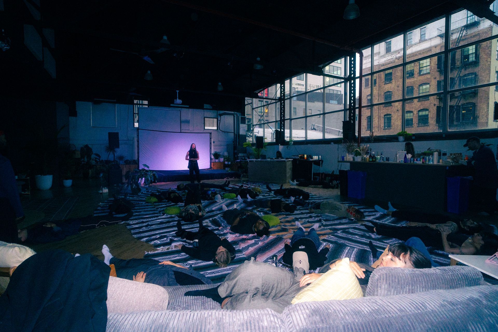 People lying down on mats, a presentation on stage, in a large room with windows and couches.
