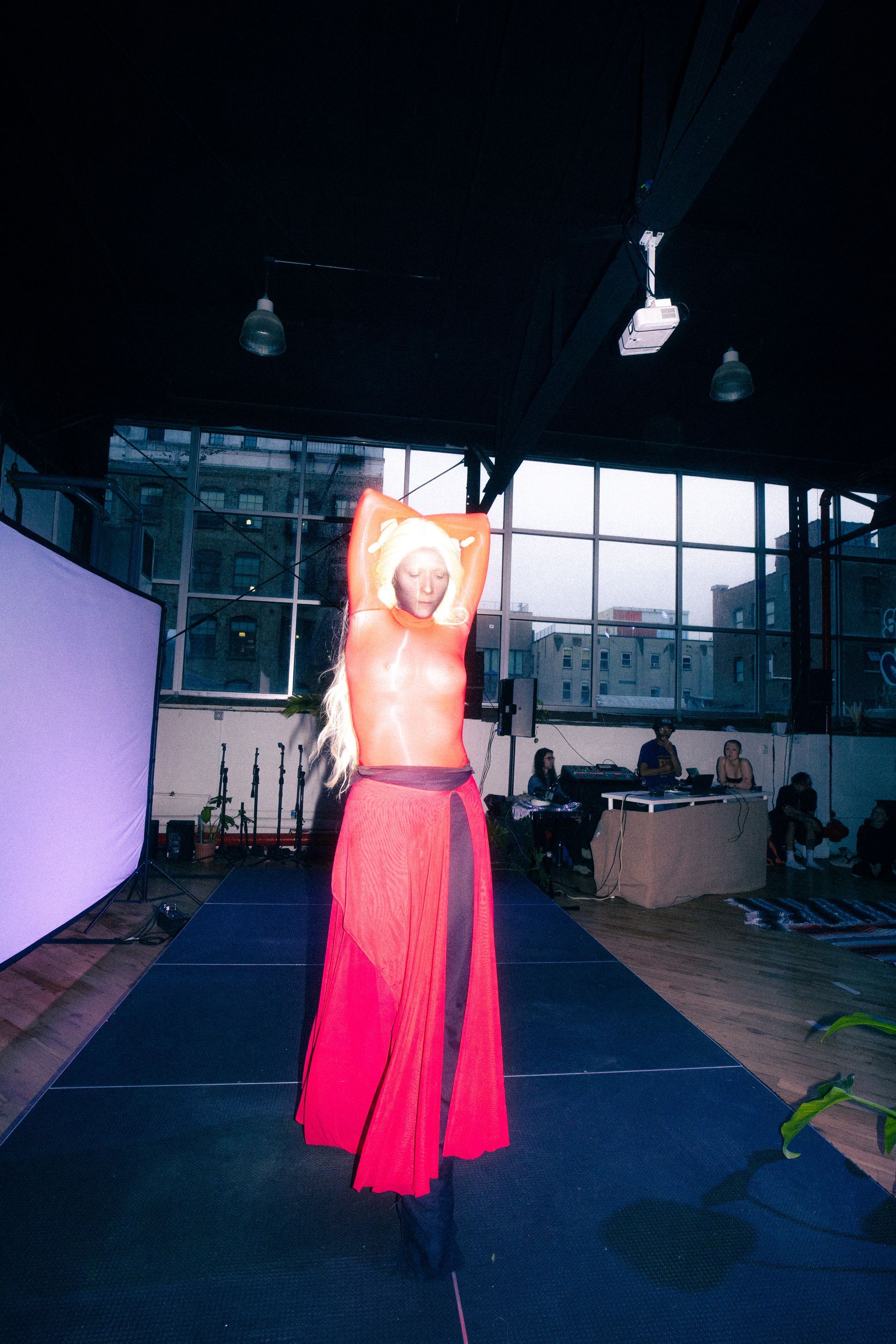 Blurred figure in red skirt and top on a runway, arms raised. Low light indoors.