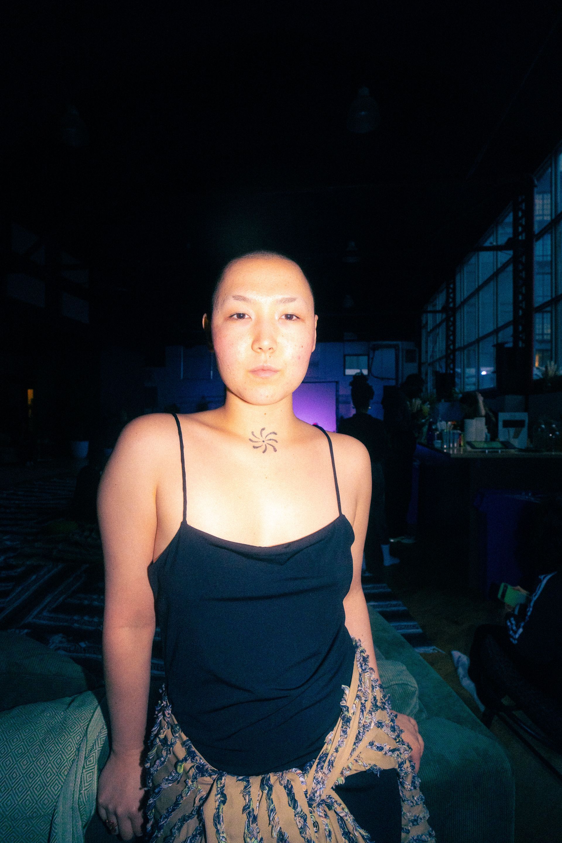 A person with a shaved head in a black tank top and patterned shawl stands indoors with dim lighting.