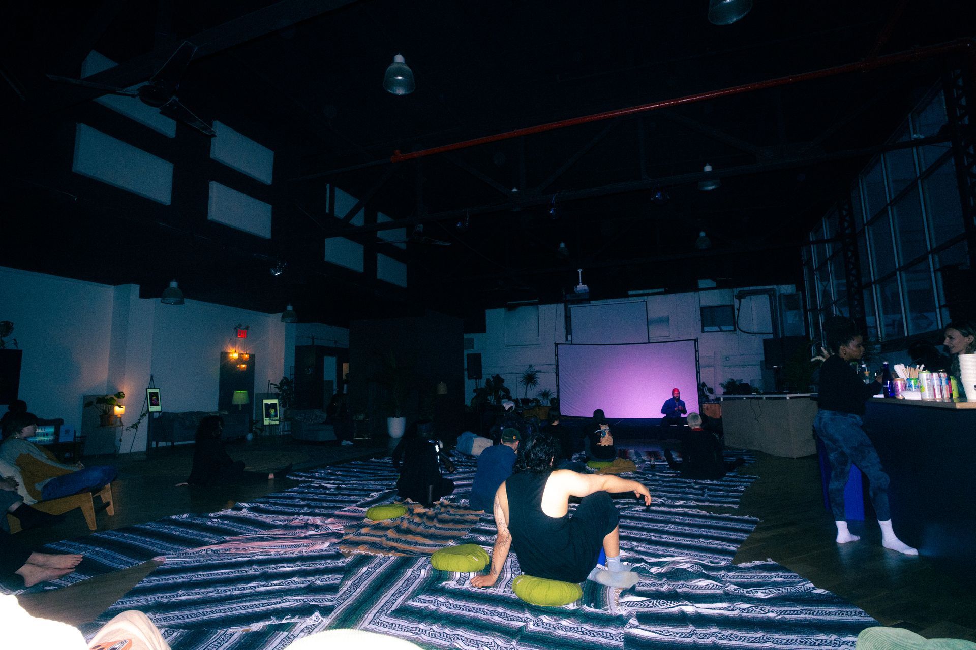 People sit on mats in a dimly lit warehouse space, possibly for a meditation or yoga class.