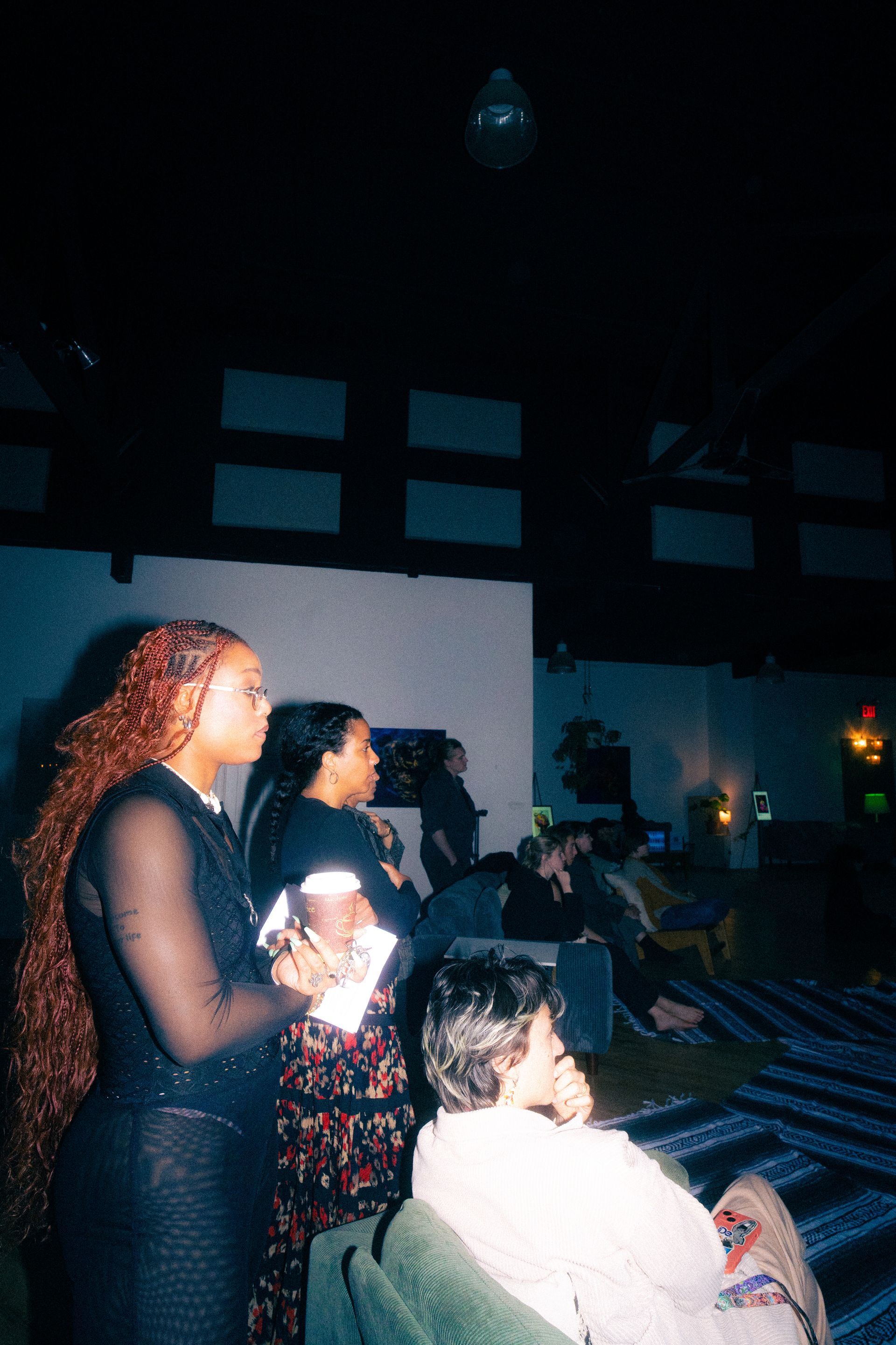 People in a dimly lit room, some standing and others seated, appear to be listening.